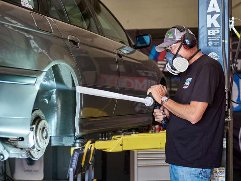 technician using dry ice blasting on the underside of a car while wearing protective gear in an auto shop