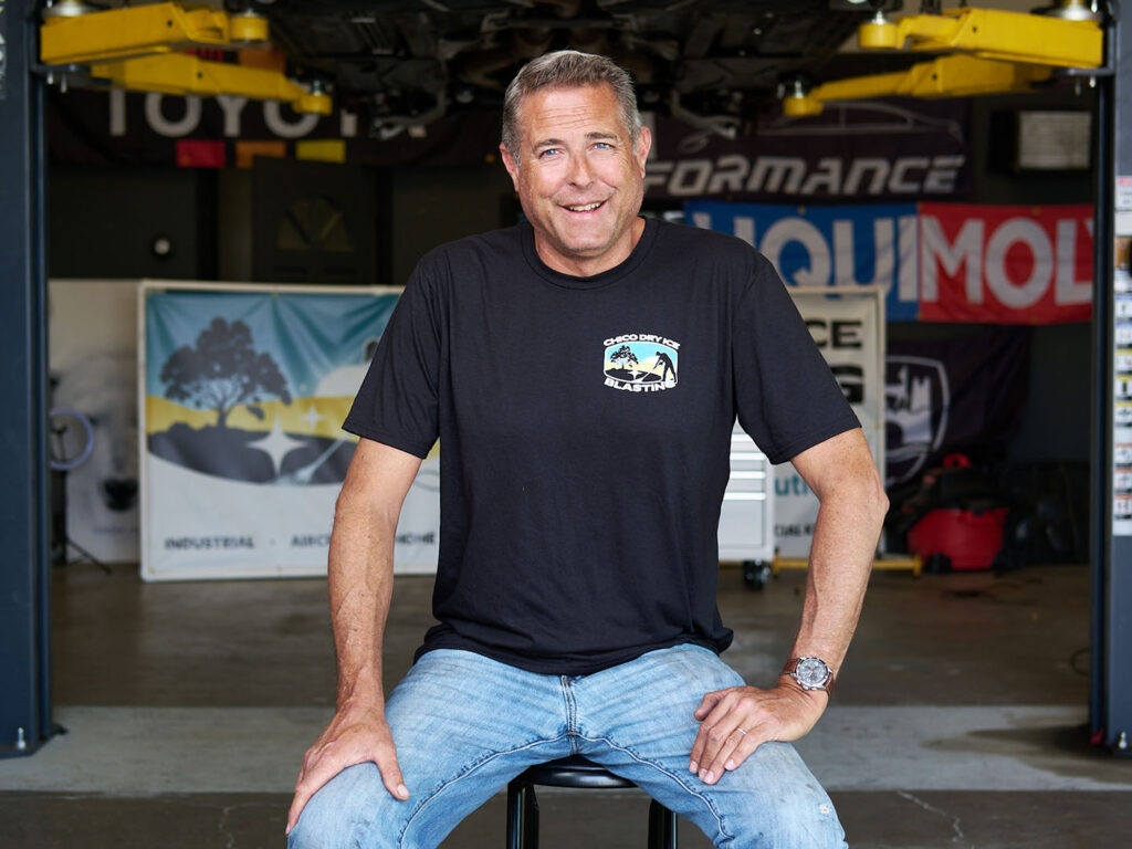 smiling man wearing a Chico Dry Ice Blasting shirt, seated in a garage workshop with banners in the background
