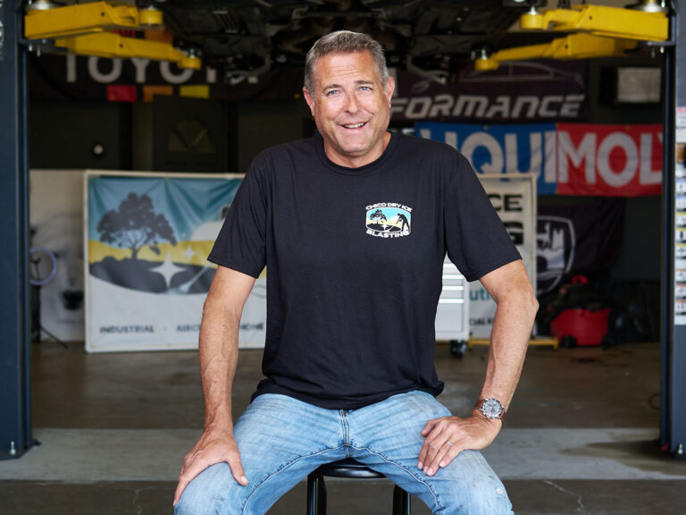 smiling man wearing a Chico Dry Ice Blasting shirt, seated in a garage workshop with banners in the background